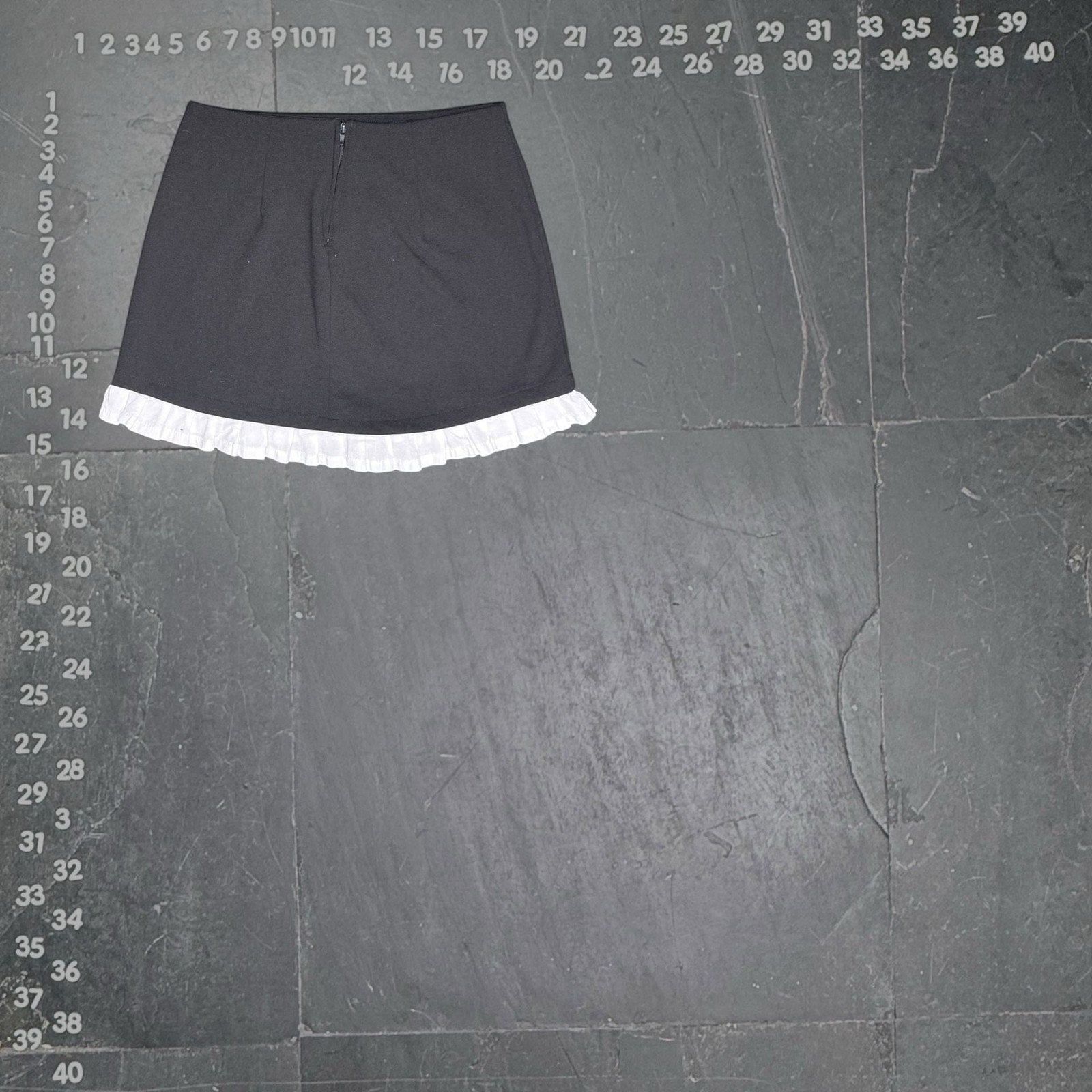 The Room Collective black mini skirt with white ruffled hem. High-waisted, slim fit, polyester/spandex blend with cotton ruffle. Back zipper.