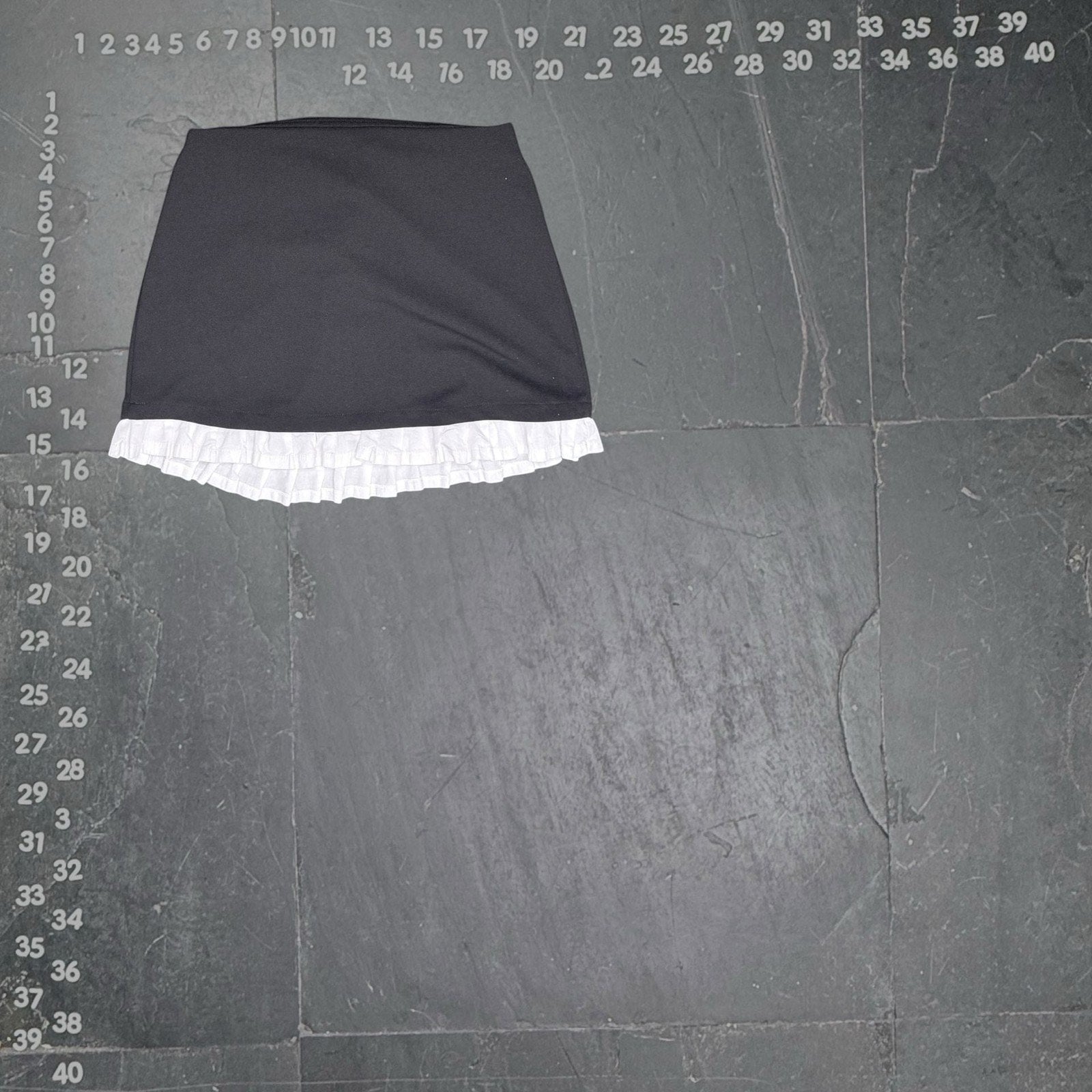 The Room Collective black mini skirt with white ruffled hem. High-waisted, slim fit, polyester/spandex blend with cotton ruffle. Back zipper.