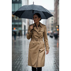 The Room Collective Khaki Tan double-breasted belted trench coat with epaulettes and belted cuffs, worn by a woman holding an umbrella in the rain.
